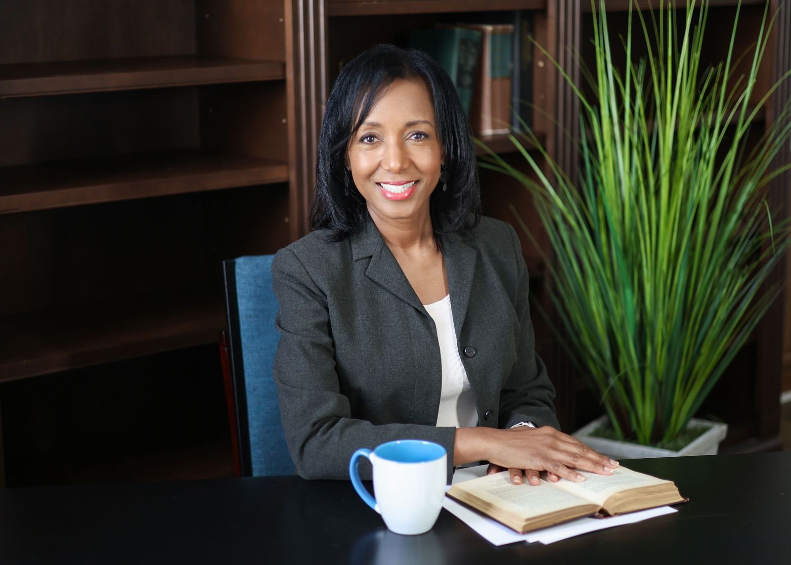 Dr. Anna-Maria Rivas McGowan smiling at a desk in a dark gray blazer, one hand resting on an open book beside a blue and white mug