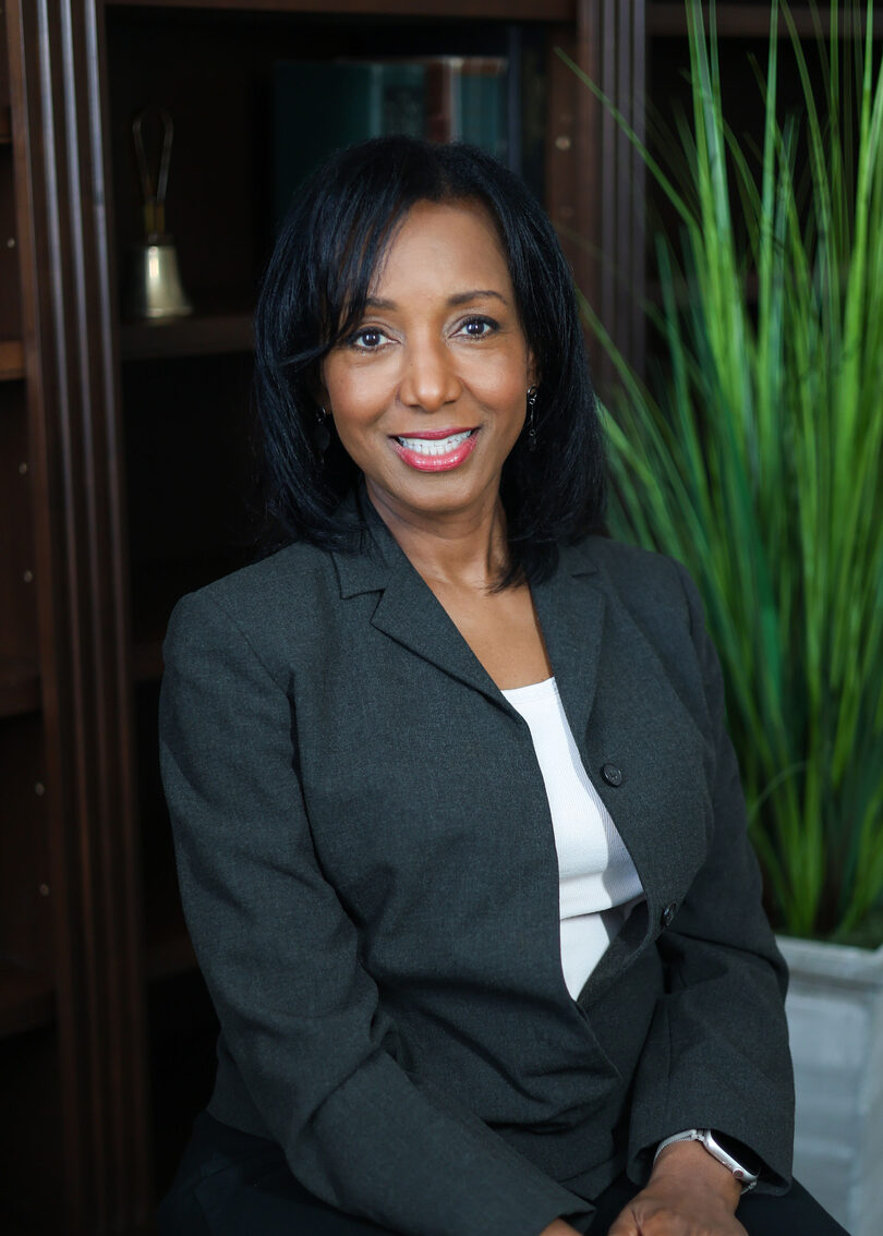 Dr. Anna-Maria Rivas McGowan in a dark gray blazer smiling in front of a wooden bookcase and a green plant