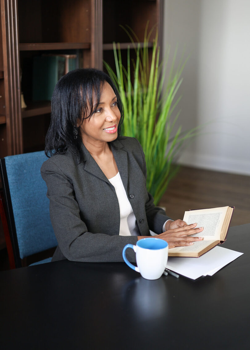 Dr. Anna-Maria Rivas McGowan seated at a table in a dark gray blazer, smiling, one hand on an open book beside a blue and white mug